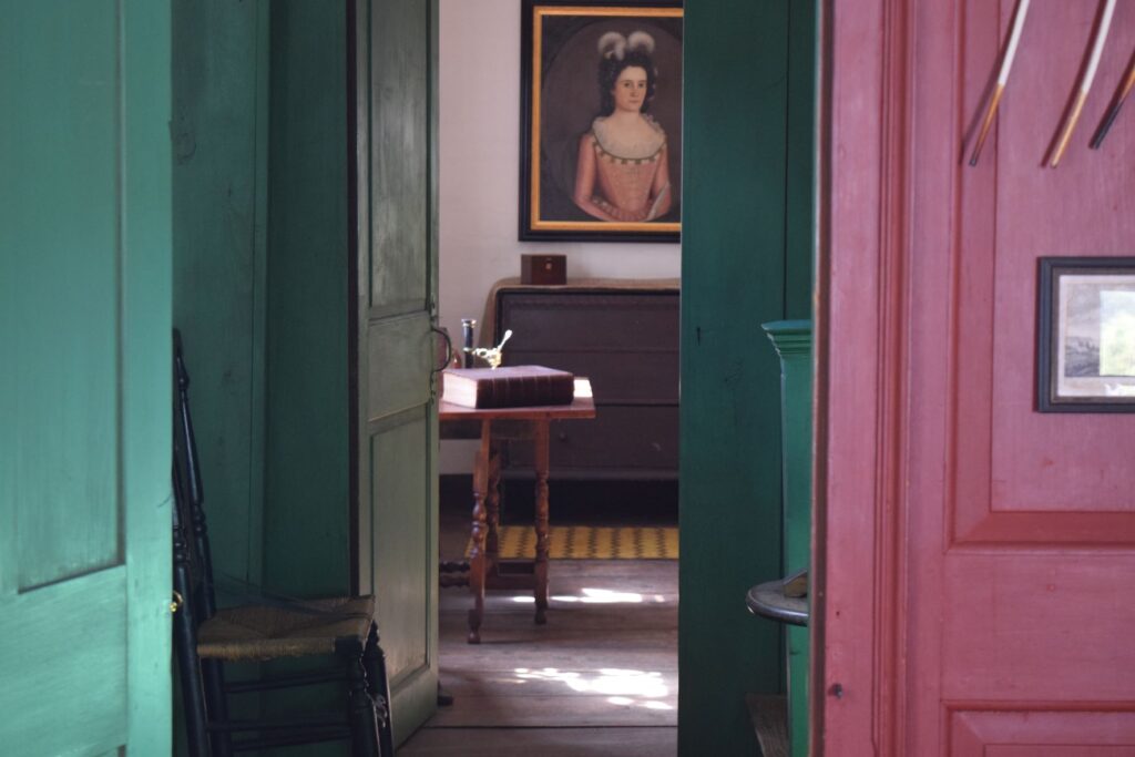 Image is taken looking into the Bidwell House Museum Parlor from the Dining Room, with the portrait of Mary Gray Bidwell on the wall. Dining Room wall is red, hallway wall is green. In the parlor you can see a desk with a book and a 3 drawer cabinet beneath the painting